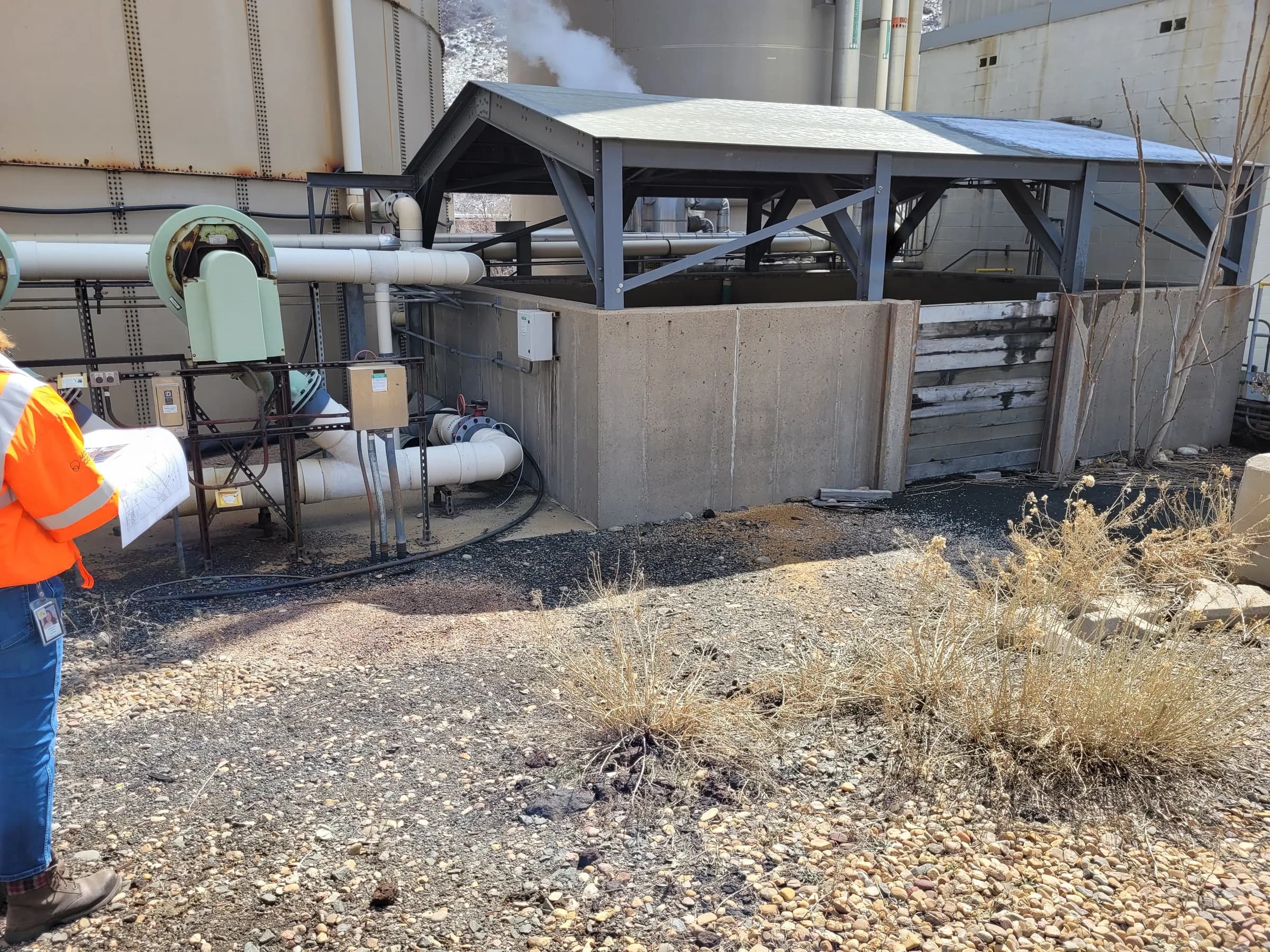 An engineer in a hi-vis jacket reviews plan sheets beside a covered concrete basin, white process piping and large tanks at an industrial plant.